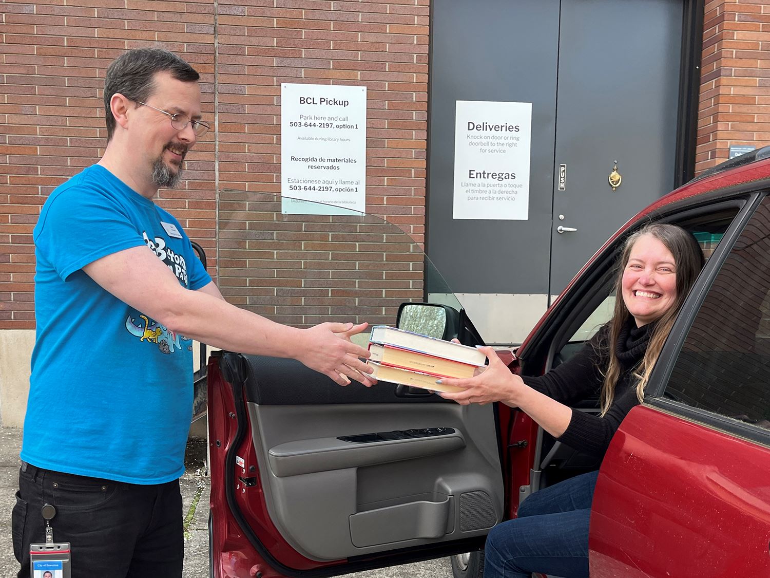 Photo of a woman sitting in a car and smiling while a man hands her a stack of books.