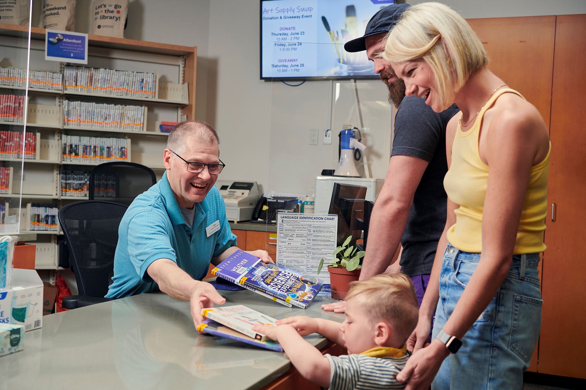 Photo of a man with glasses sitting at a desk checking out books for a family with a child.