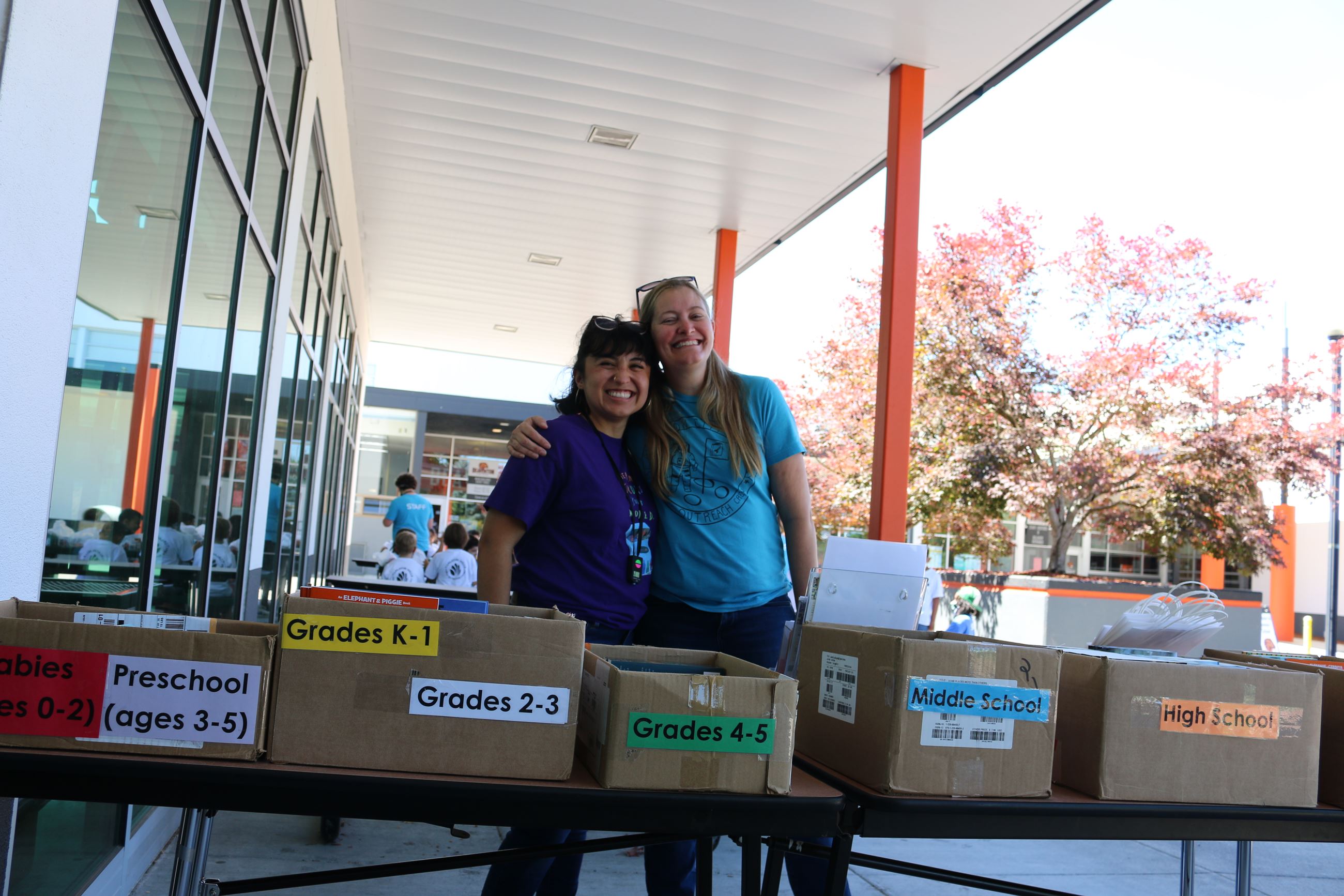Photo of two smiling women standing at a table with boxes of books labeled for different grades.